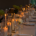 Elegant wedding reception table with glowing pillar candles in glass hurricanes, crystal glassware, and green floral arrangements