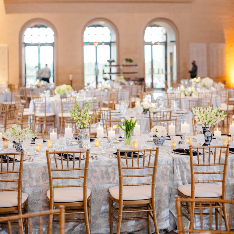 Elegant ballroom reception with gold chiavari chairs, patterned linens, white floral centerpieces, and arched windows