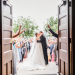 Newlywed couple kissing while walking through church doors as guests celebrate with ribbon wands