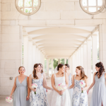Bride in white gown with bridesmaids in floral and gray dresses holding bouquets under white covered walkway