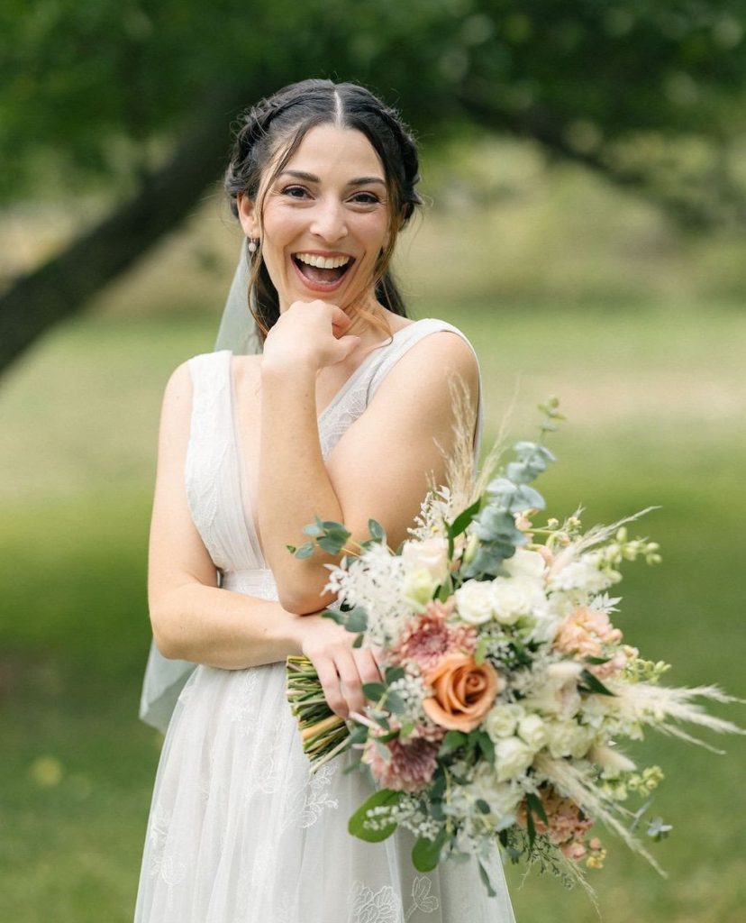 Joyful bride laughing in white lace gown and veil, holding romantic bouquet of peach roses and eucalyptus