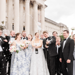 Newlyweds and wedding party celebrating with champagne spray in front of classical white columned building