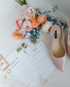 Wedding day flatlay with bridal bouquet in blush and blue tones, pink heels, invitation suite, and rings