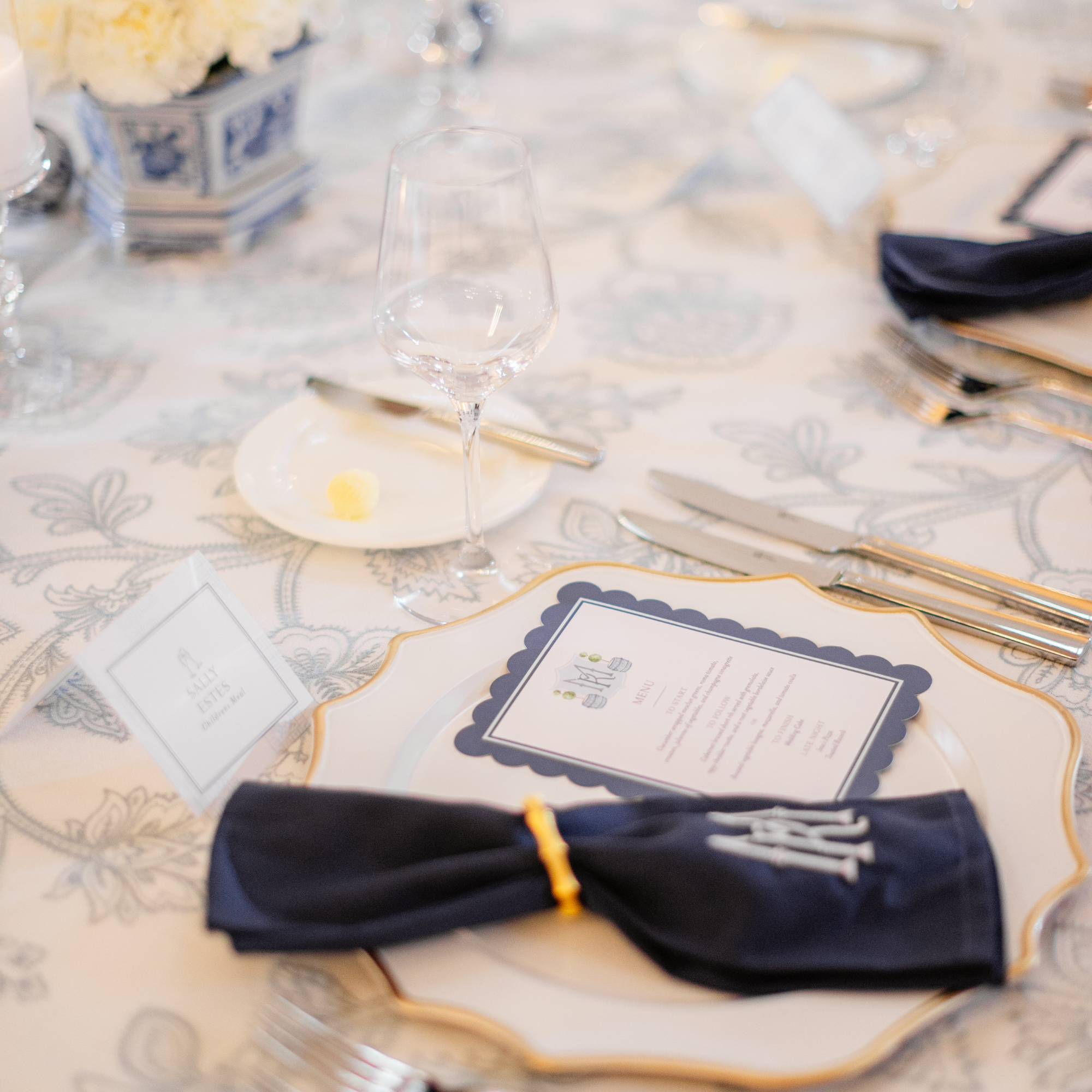 Elegant place setting with navy napkin, scalloped menu card, gold flatware, and patterned tablecloth