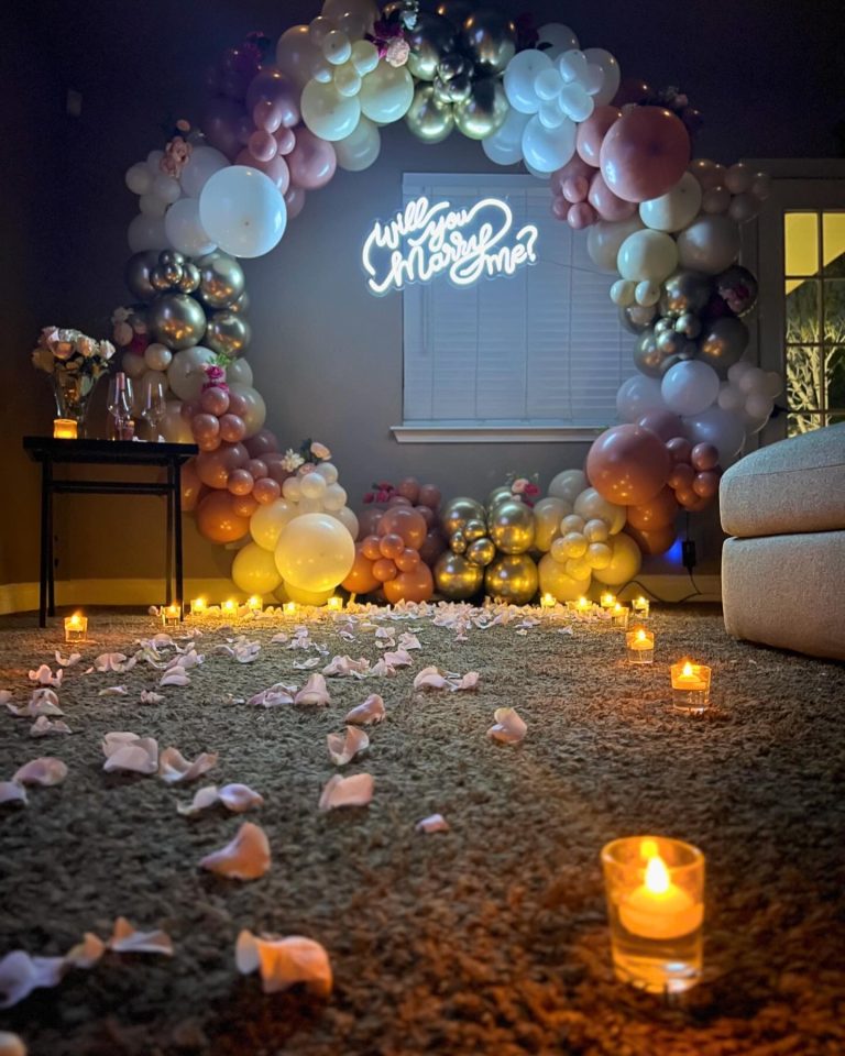 Romantic marriage proposal setup with pastel balloon arch, "Will you Marry Me?" neon sign, rose petals, and candles