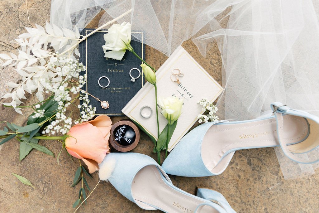 Wedding details flat lay featuring blue heels, rings, vows booklet, white roses, and veil