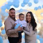 Family of three posing beneath blue and white balloon arch