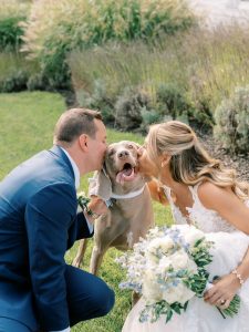Bride and groom kissing their happy dog wearing a bow tie, holding white and blue bouquet outdoors