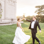Bride and groom walking hand-in-hand on lawn in front of white classical building at sunset