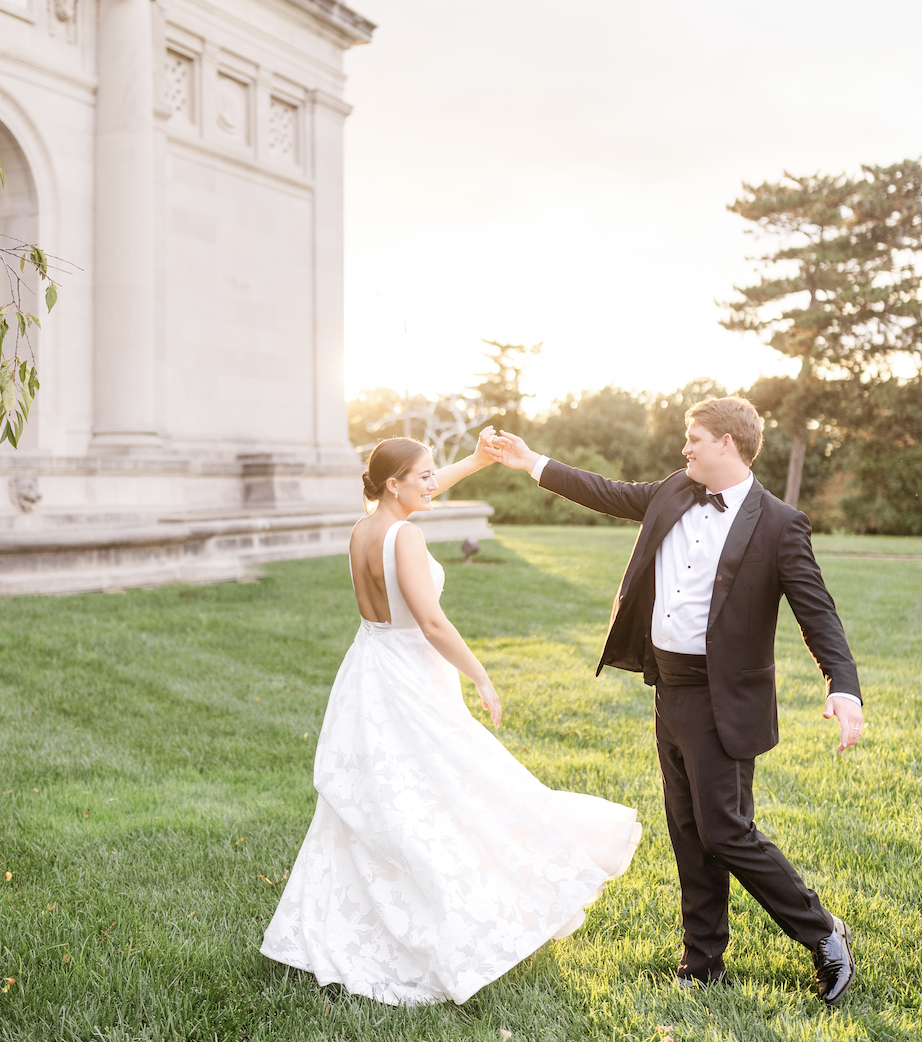 Bride and groom walking hand-in-hand on lawn in front of white classical building at sunset