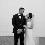 Newlywed couple embracing against white wall, groom in black tuxedo, bride in white strapless gown with veil