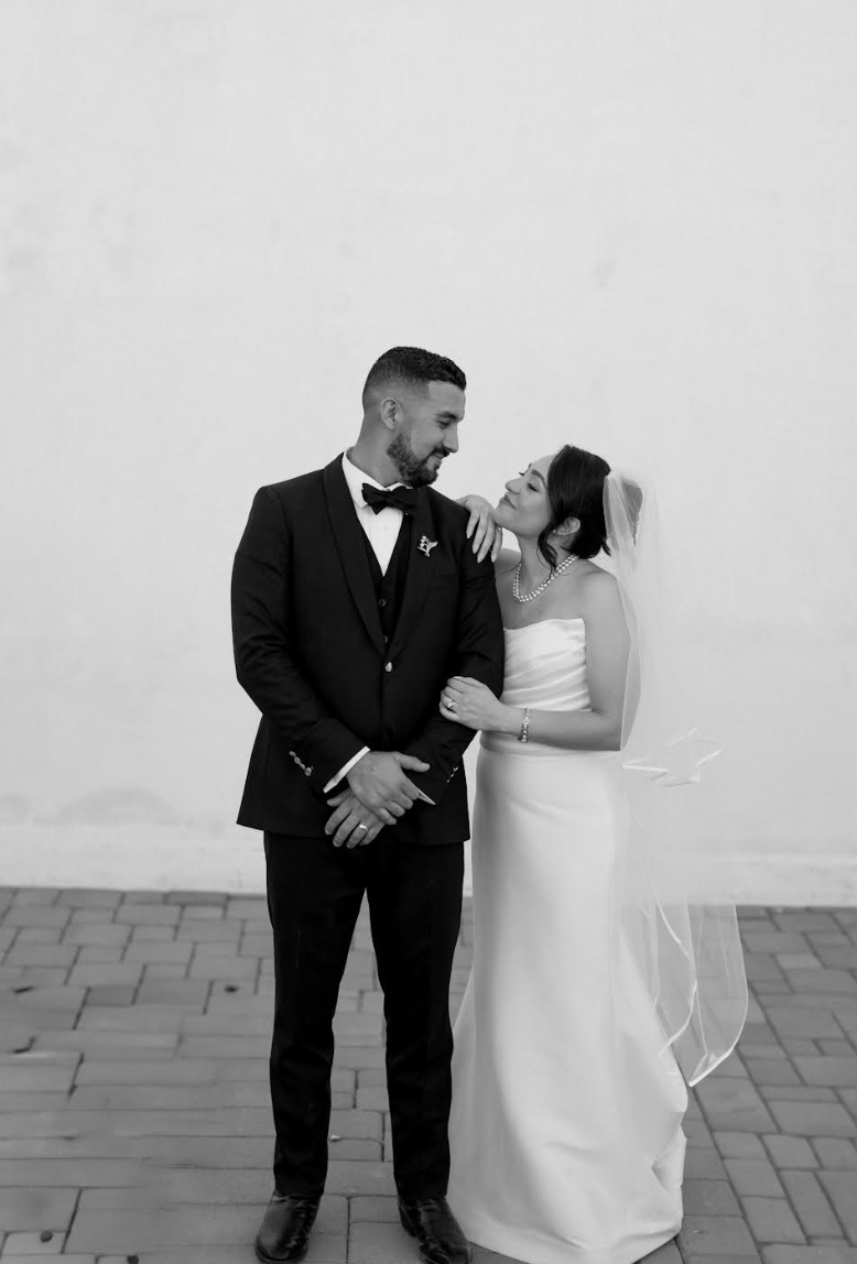 Newlywed couple embracing against white wall, groom in black tuxedo, bride in white strapless gown with veil