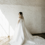 Bride in flowing gown with pearl-embellished veil poses against white brick wall in natural light