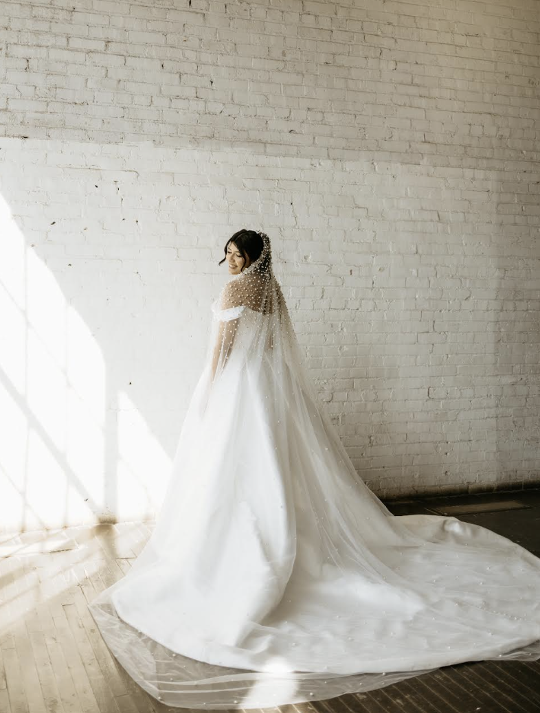 Bride in flowing gown with pearl-embellished veil poses against white brick wall in natural light