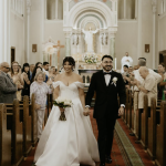 Newlyweds walking down church aisle as guests applaud in ornate white cathedral