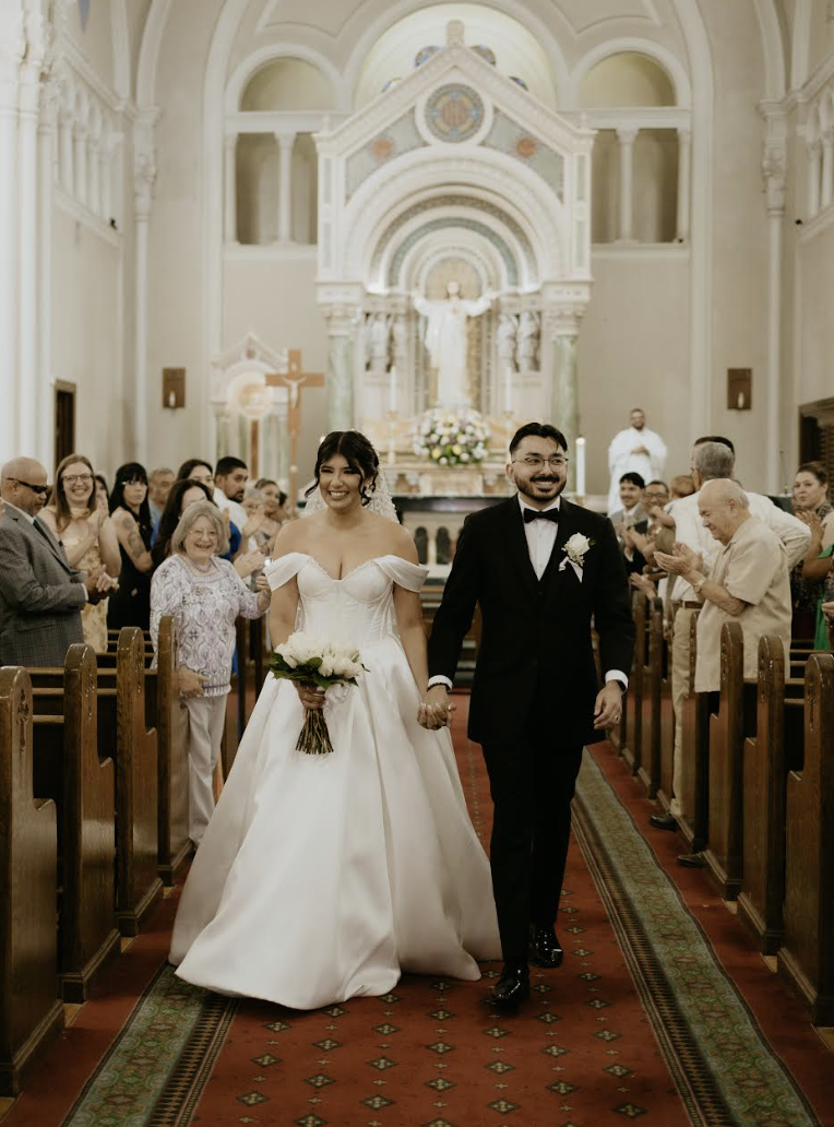 Newlyweds walking down church aisle as guests applaud in ornate white cathedral