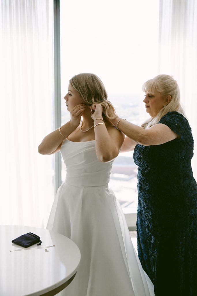 Bride Leah adjusts her earrings in an elegant white gown while her mother assists with wedding preparations