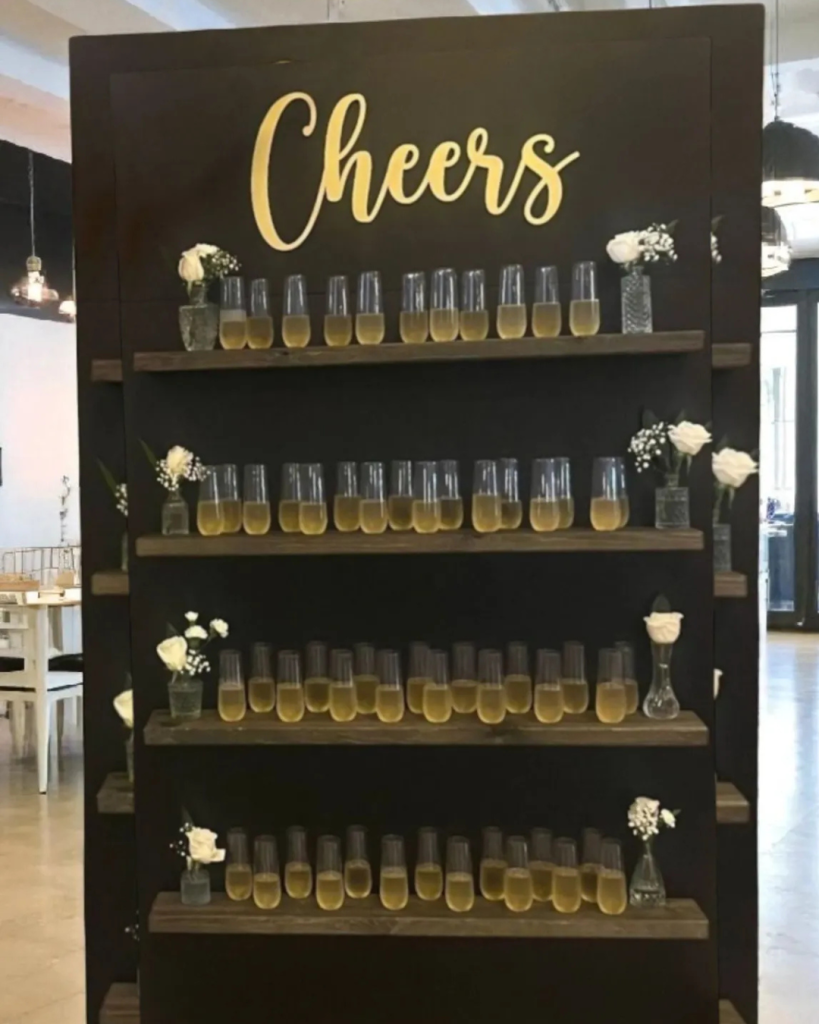 Black champagne wall with gold "Cheers" lettering displaying stemless flutes and white rose accents on wooden shelves