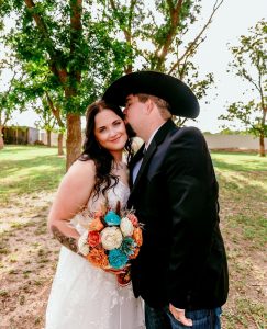 Groom in cowboy hat kisses bride's cheek as she holds turquoise and orange wood flower bouquet in outdoor garden setting