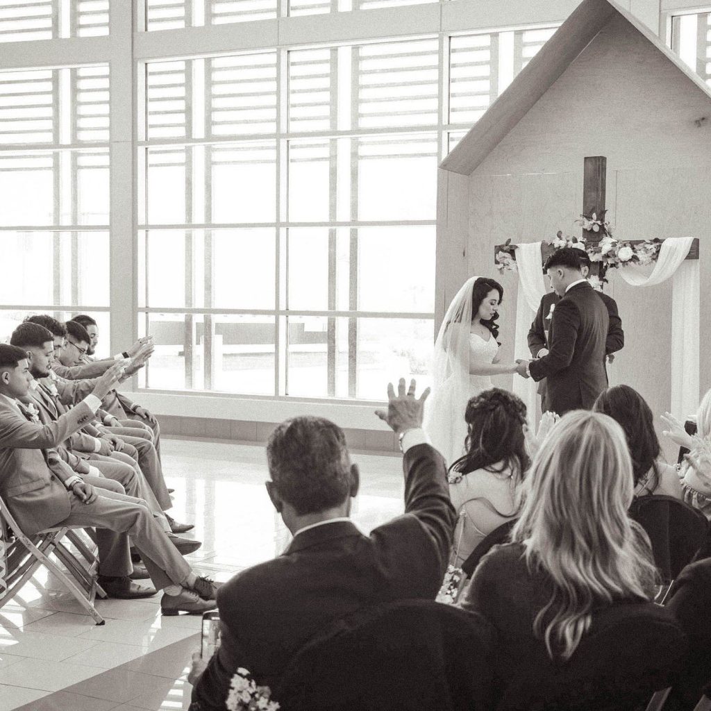 Bride and groom exchanging vows at intimate indoor ceremony with guests seated in rows and wooden chapel backdrop