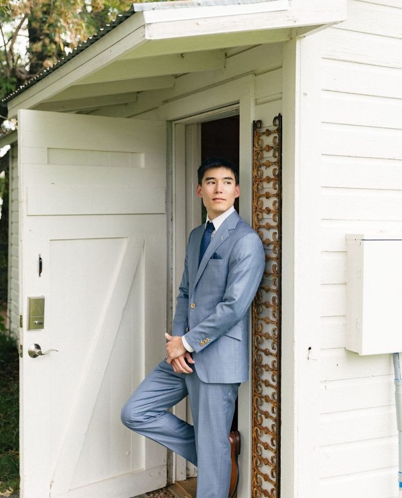 Groom in light blue suit posing confidently in doorway of white rustic barn at Laporte wedding venue