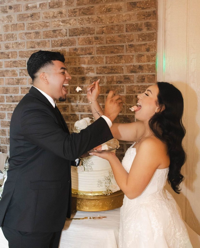 Newlyweds playfully feeding each other wedding cake against exposed brick wall