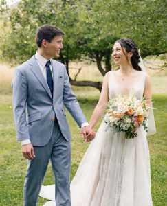 Newlywed couple holding hands and laughing together in outdoor garden setting with bride holding peach and cream rose bouquet