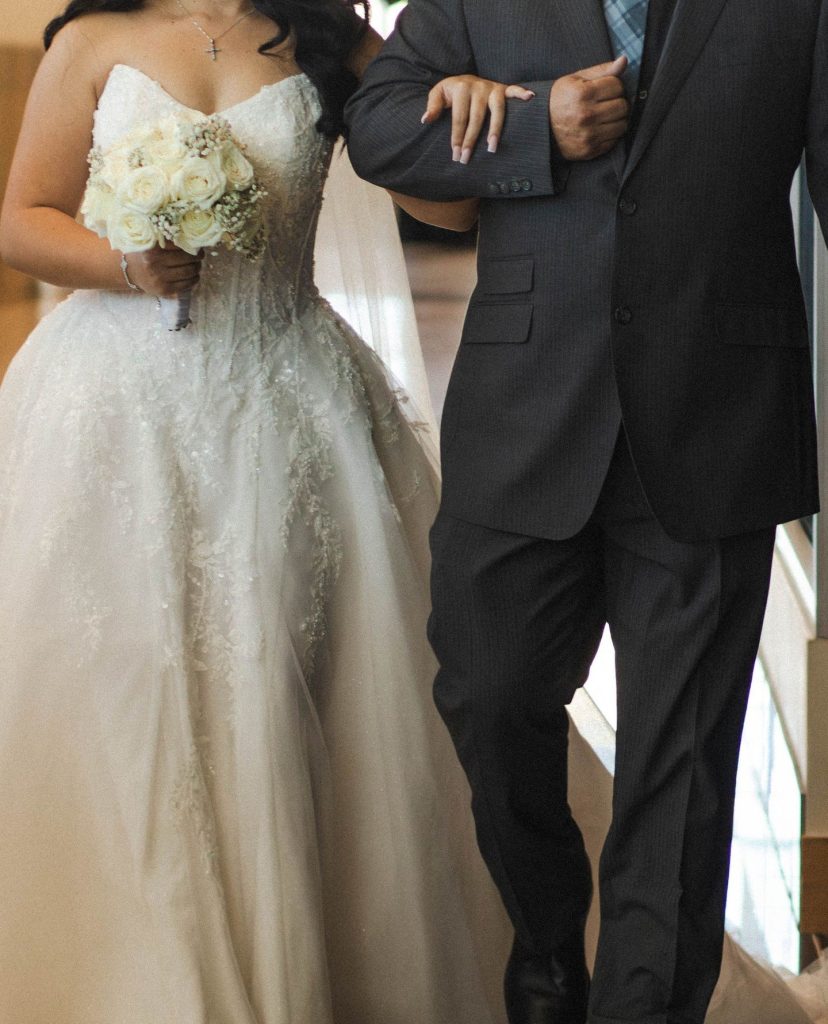 Bride in sparkly ballgown holding white rose bouquet walks arm-in-arm with groom in charcoal suit