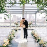 Bride and groom kissing at modern Cleveland wedding ceremony with floor-to-ceiling windows, clear ghost chairs, and lush floral arrangements