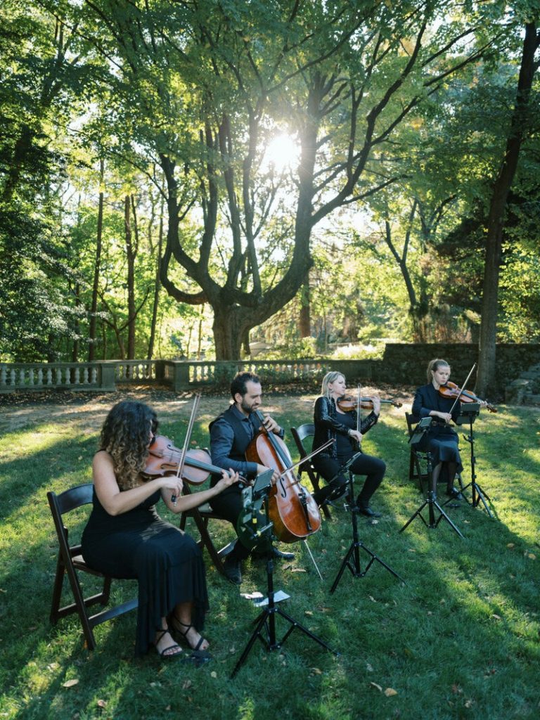 String quartet performs outdoors in a sunlit garden setting at a wedding ceremony