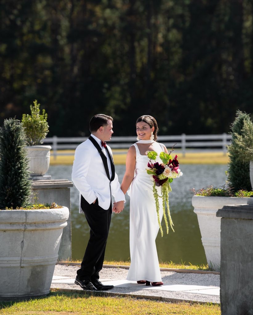 Couple standing hand-in-hand on lakeside path with bride holding cascading burgundy and green bouquet