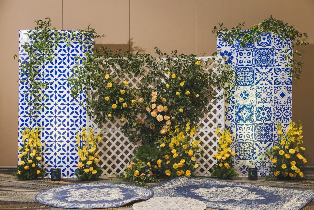 Mediterranean-inspired wedding backdrop featuring blue patterned tile panels, yellow floral arrangements, and lattice with climbing greenery