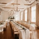 Elegant ballroom with crystal chandeliers, draped white ceiling fabric, long banquet tables with gold chiavari chairs and greenery garland runners