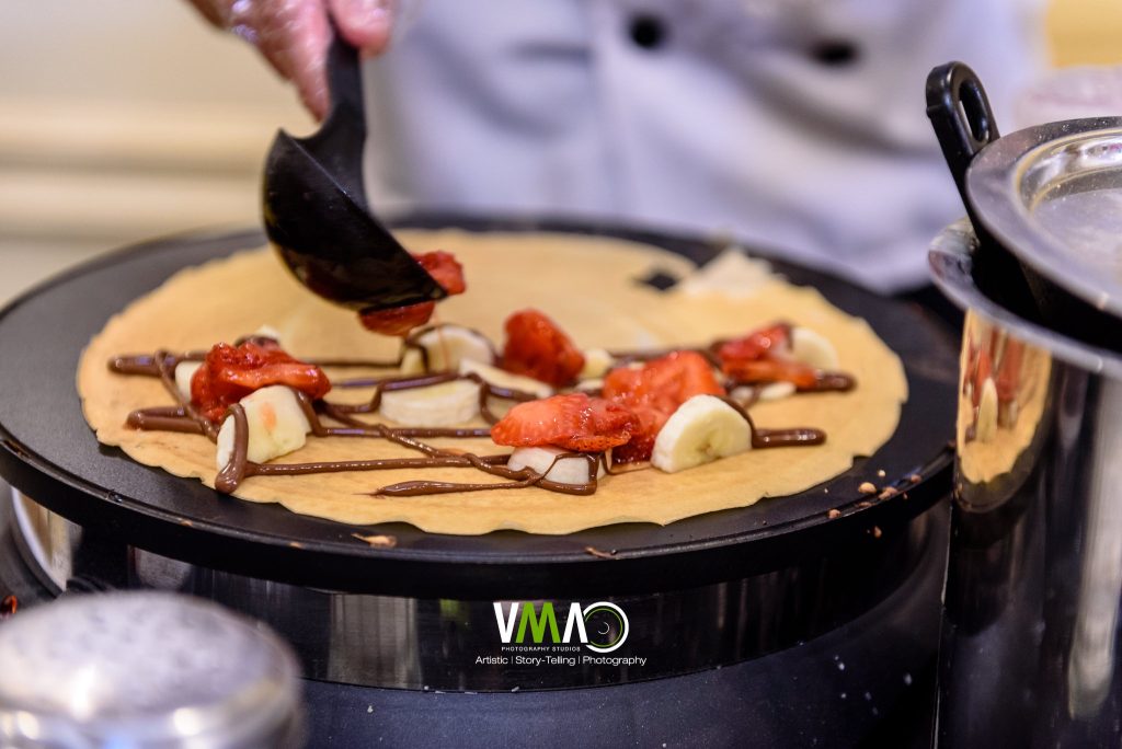 Chef preparing crepe with strawberries, banana, and chocolate on griddle at wedding catering station