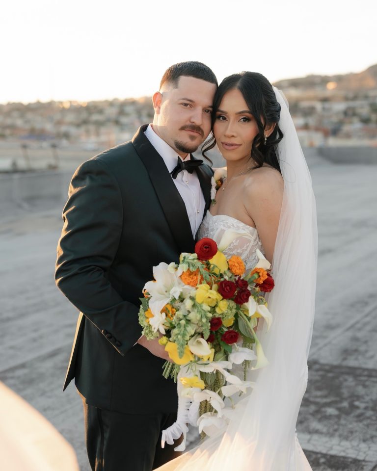 Bride and groom in formal wedding attire posing at sunset with colorful bouquet featuring red, yellow, and white roses