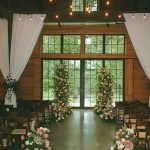 Rustic barn wedding ceremony space with draped white curtains, string lights, tall pink and peach floral columns flanking glass doors, and wooden chairs lining the aisle