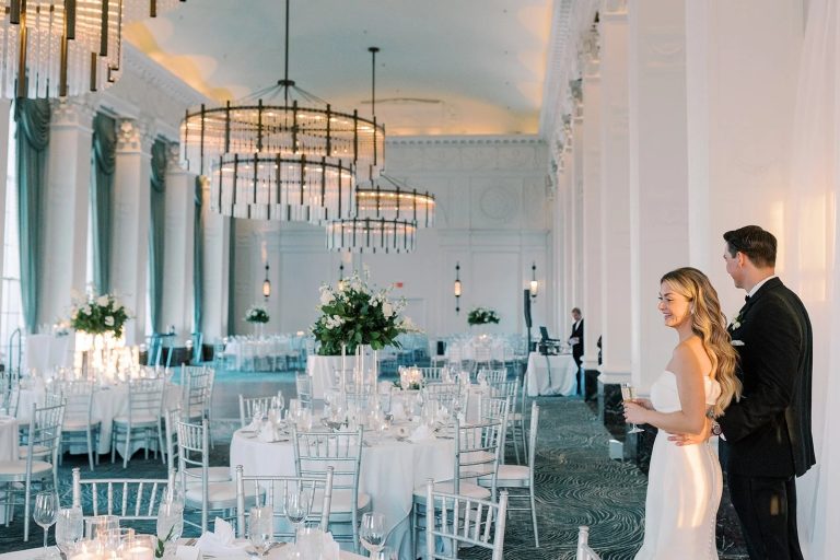 Bride and groom admiring elegant ballroom with crystal chandeliers, white chiavari chairs, and tall floral centerpieces
