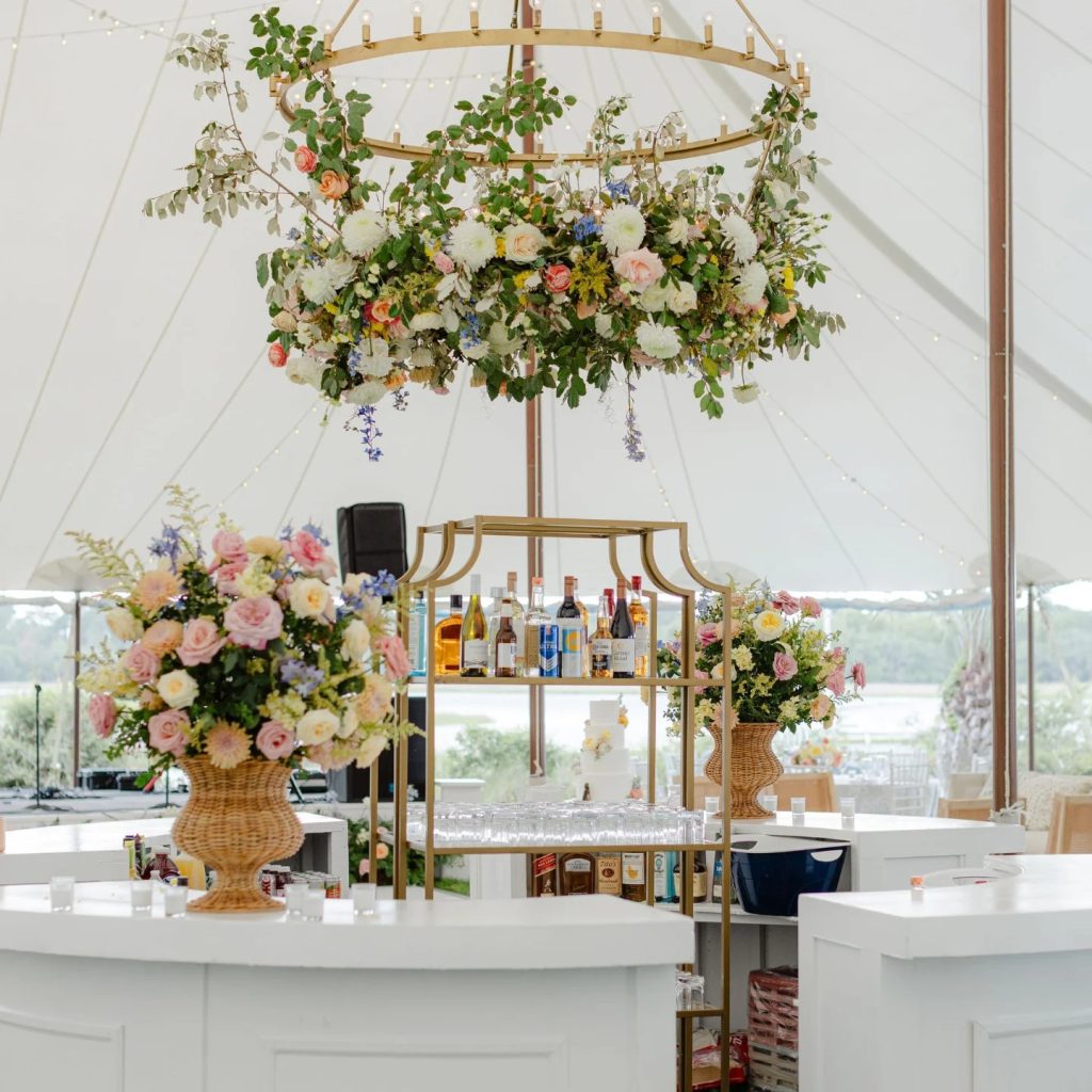 Elegant white bar setup under tent with gold chandelier adorned with colorful flowers, gold bar cart, and floral arrangements in wicker urns