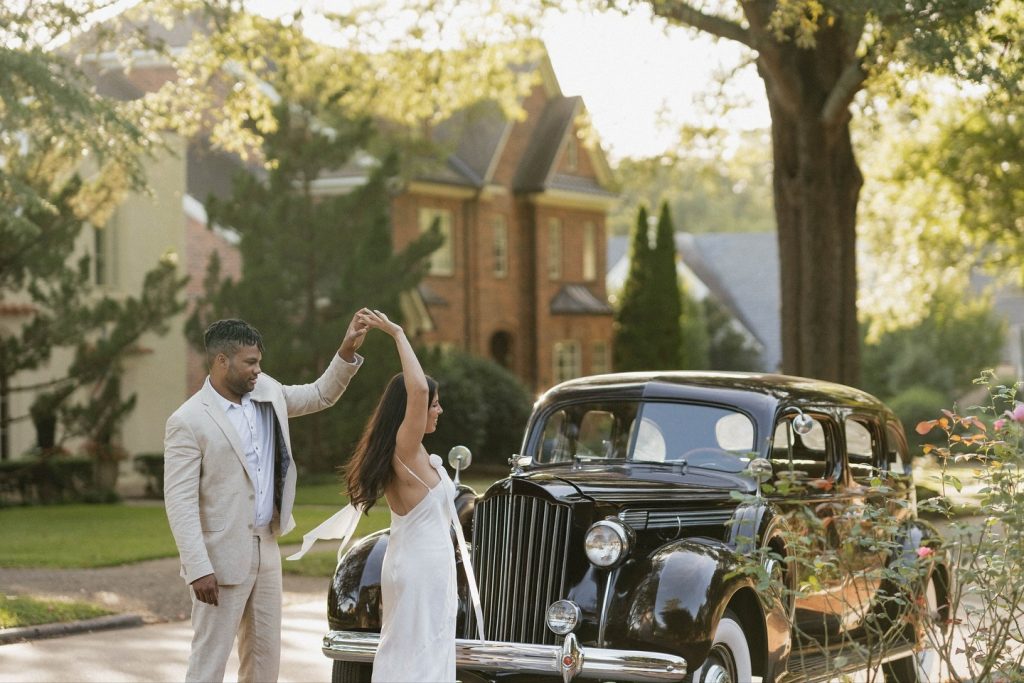Newlyweds walking beside vintage black classic car on tree-lined driveway