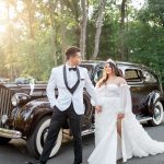 Bride and groom posing beside vintage brown classic car in outdoor wooded setting