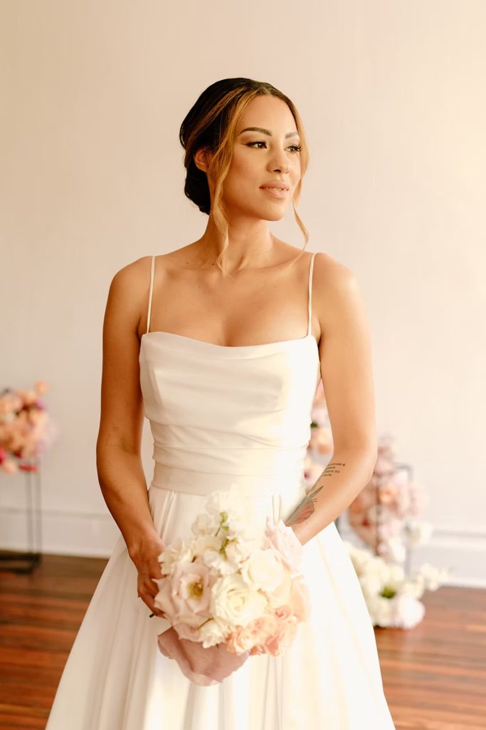 Bride in white gown with elegant updo holds blush and ivory bouquet in preparation room
