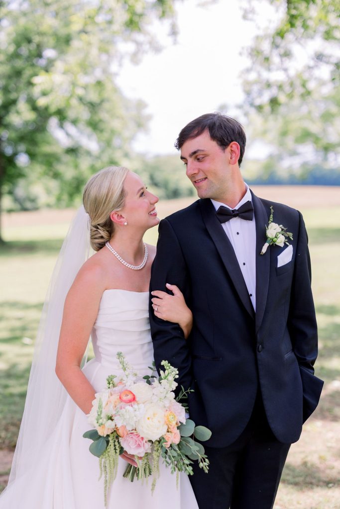 Bride in strapless gown and pearl necklace gazes at groom in classic black tuxedo outdoors