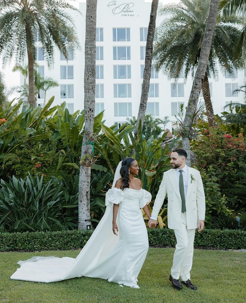 Bride in off-shoulder gown with cathedral train and groom in cream suit walk hand-in-hand across tropical hotel grounds