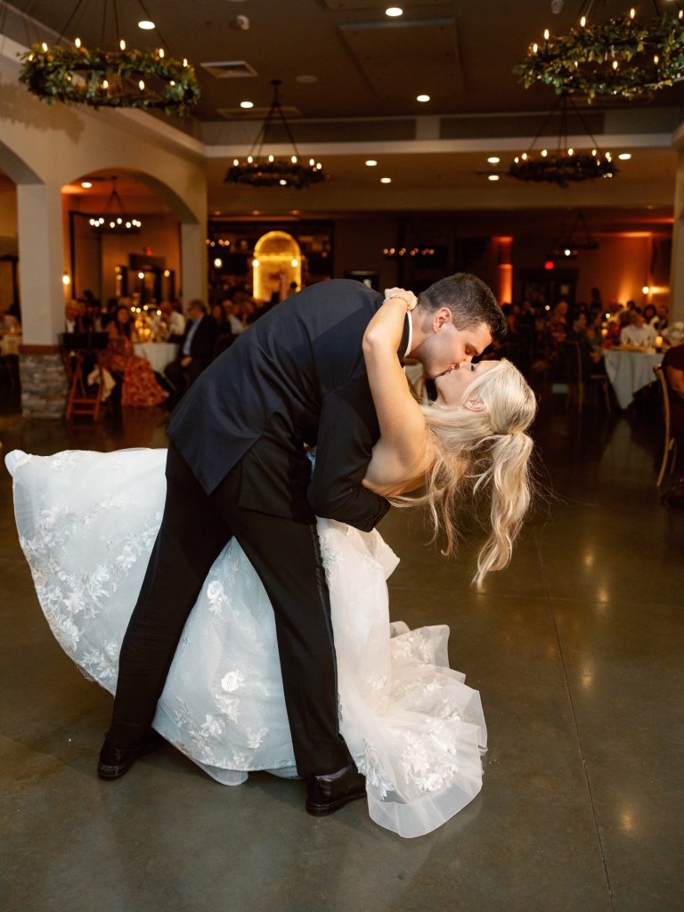Bride and groom sharing dramatic first dance kiss in warmly lit reception hall