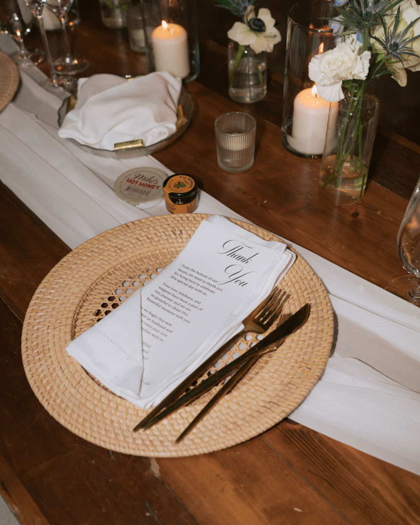 Rustic place setting with woven rattan charger, gold flatware, menu card, and white flowers on dark wood table