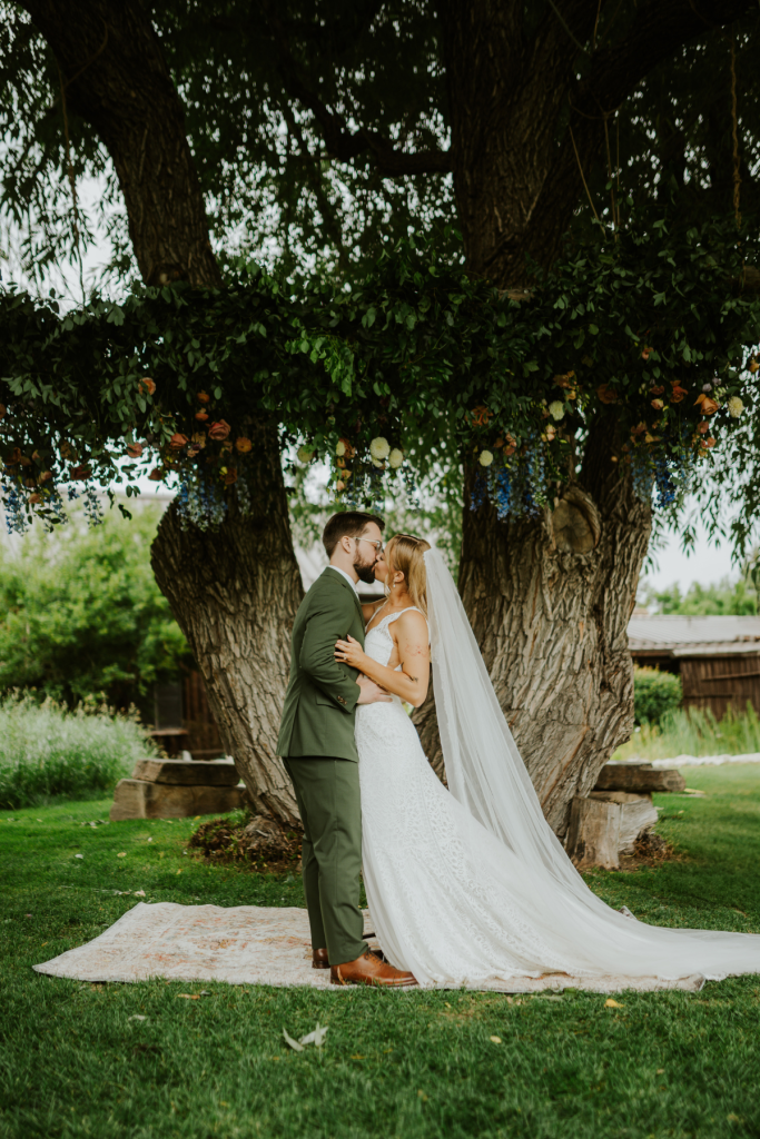 Mallory and Andrew embrace beneath a floral-adorned tree at Spruce Mountain Ranch, her cathedral veil flowing across the grass