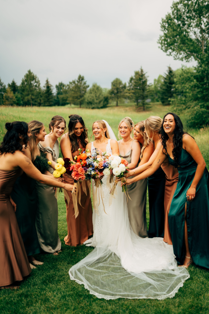 Bride Mallory with bridesmaids in emerald and rust gowns admiring colorful bouquets on mountain meadow