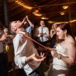 Groom performs limbo at wedding reception while bride holds pole and guests cheer at rustic barn venue