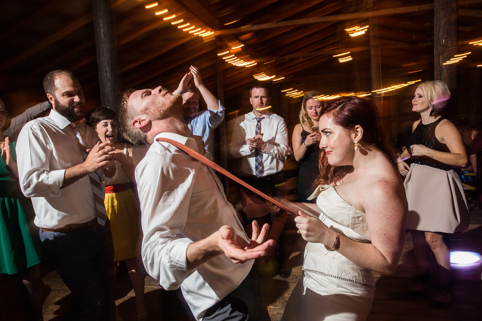 Groom performs limbo at wedding reception while bride holds pole and guests cheer at rustic barn venue