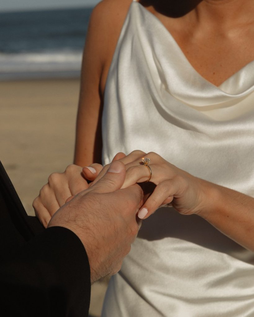 Close-up of bride’s engagement ring as groom holds her hand on beach at intimate Addy Sea wedding in Bethany Beach, Delaware.